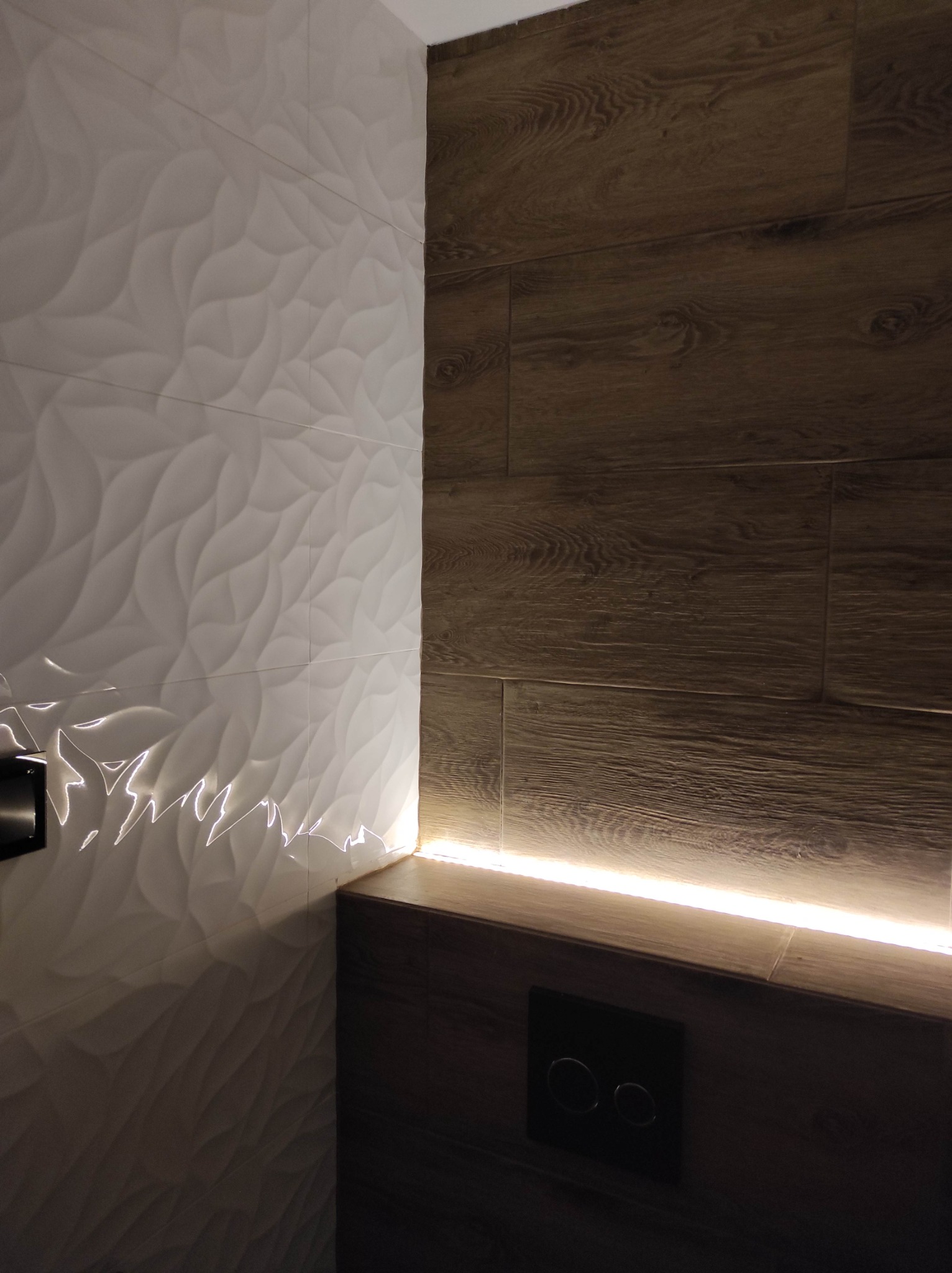 Nowoczesna łazienka z białymi płytkami 3D i drewnopodobnymi panelami, podświetlenie LED wzdłuż krawędzi, czarny panel sterowania spłuczką.