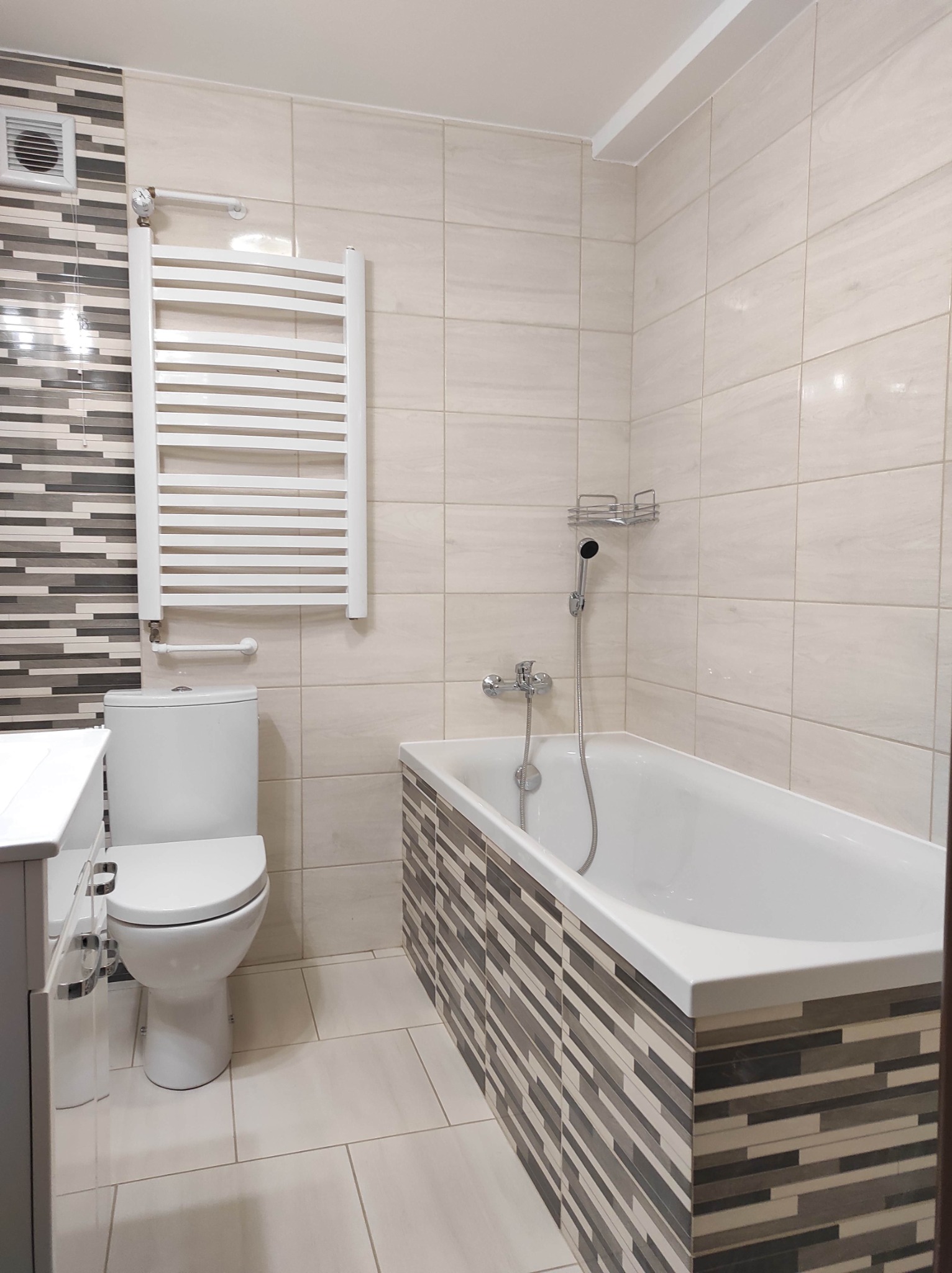 Jasna łazienka po remoncie: beżowe płytki na ścianach i podłodze, wanna obudowana mozaiką w odcieniach szarości i beżu, biała toaleta, grzejnik drabinkowy i umywalka z szafką.