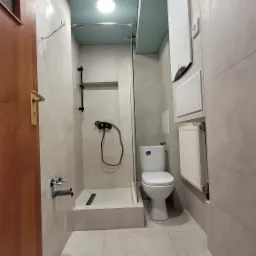 Wykończona łazienka z kabiną prysznicową typu walk-in, wiszącą toaletą, grzejnikiem drabinkowym i bojlerem, wykończona płytkami w odcieniach szarości, z surowym, pomalowanym na niebiesko sufitem...