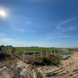 Montaż białego ogrodzenia panelowego na polu, widoczny minikoparka i pracownik w pomarańczowej koszulce, błękitne niebo z lekkimi chmurami.