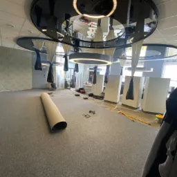 Rozwinięta rolka szarej wykładziny dywanowej na podłodze biura z widocznymi narzędziami montażowymi i elementami wystroju wnętrza, w trakcie instalacji.