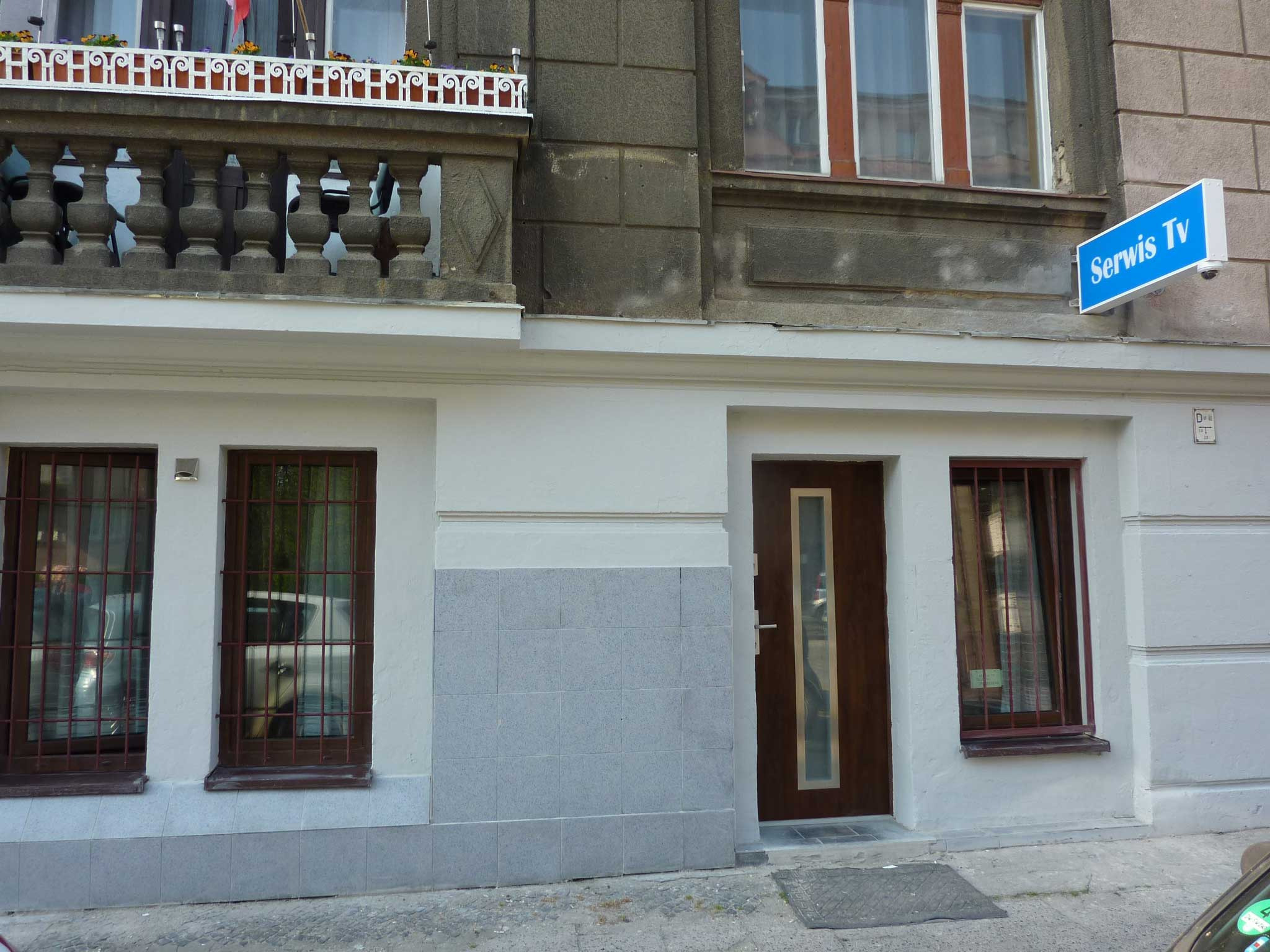 Fasada budynku z szyldem 'Serwis TV', okna z kratami i balkon z balustradą.