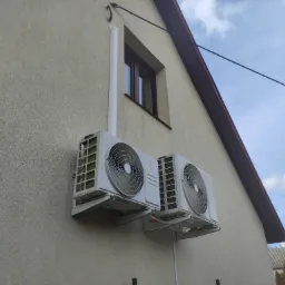 Dwa białe klimatyzatory marki Rotenso zamontowane na elewacji domu, widoczne rury odprowadzające skropliny i okno na tle błękitnego nieba.