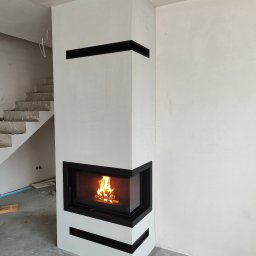 LUX-BUD Łukasz Krawczyk - Nowoczesny kominek narożny z czarnymi akcentami, w surowym wnętrzu. Widoczne niedokończone schody i betonowa podłoga. Ogień płonie w palenisku.