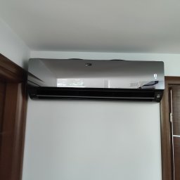 LUX-BUD Łukasz Krawczyk - Czarny klimatyzator ścienny z wyświetlaczem temperatury 18 stopni, zamontowany na białej ścianie w pomieszczeniu z drewnianą futryną drzwi.