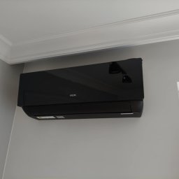 LUX-BUD Łukasz Krawczyk - Czarny klimatyzator AUX zamontowany na szarej ścianie w pomieszczeniu z listwą dekoracyjną pod sufitem i widocznym fragmentem okna.