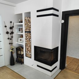 LUX-BUD Łukasz Krawczyk - Biały kominek narożny z czarnymi akcentami, obok półki z drewnem i dekoracjami. Widoczny fragment podłogi i drzwi. Nowoczesny design.