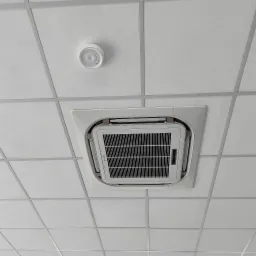 Kasetonowa klimatyzacja sufitowa wkomponowana w biały sufit podwieszany, widok z dołu. Obok czujnik dymu. Widoczne kratki wentylacyjne i panel sterowania.