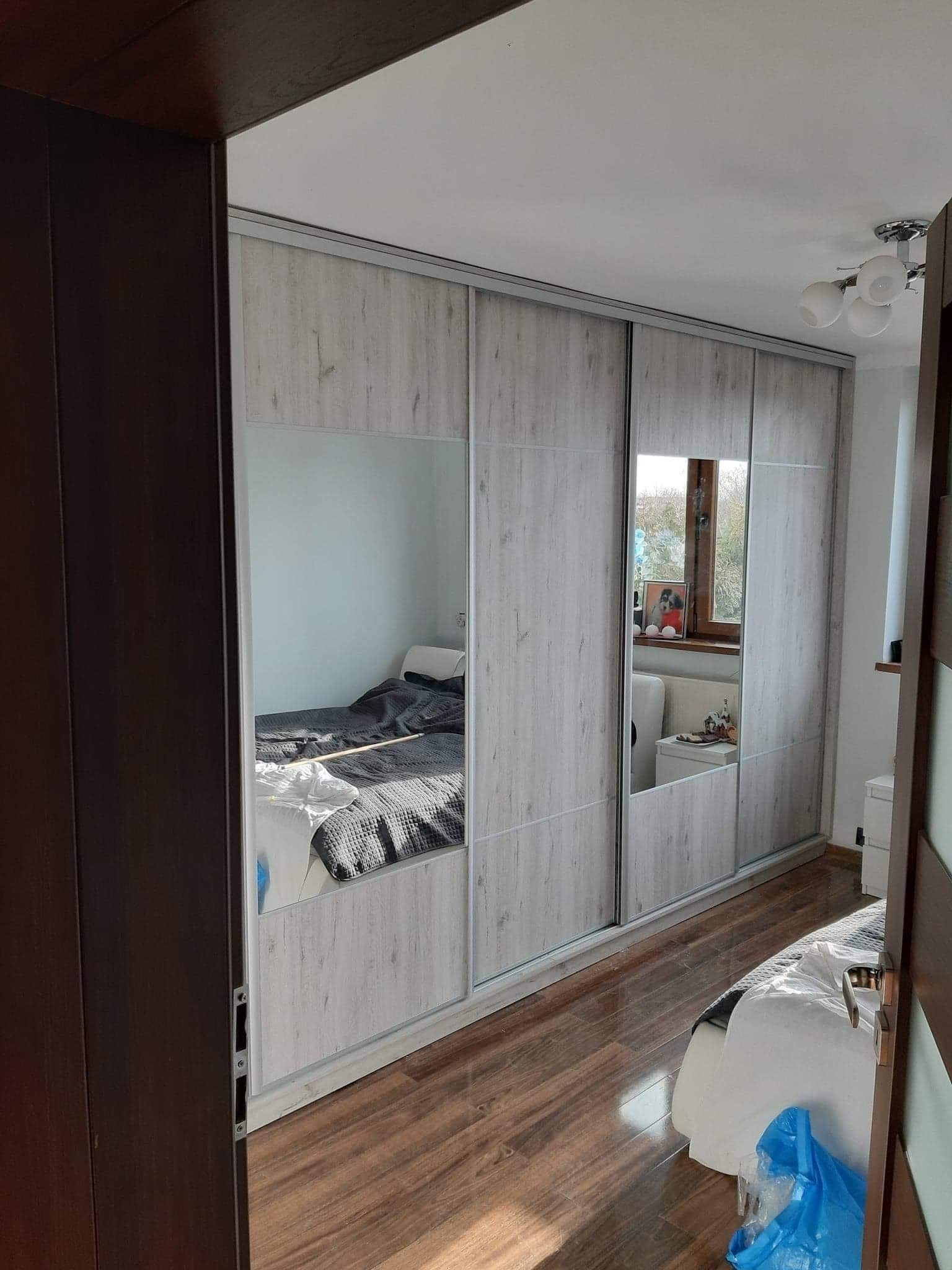 Szafa wnękowa z przesuwnymi drzwiami z lustrami, w odcieniach bieli i szarości, widok z perspektywy drzwi wejściowych do sypialni, odbicie łóżka w lustrach.