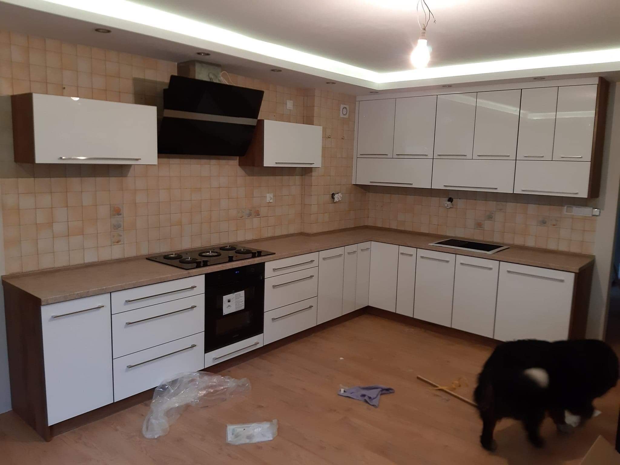 Nowoczesna kuchnia z białymi szafkami i brązowym blatem, czarny okap nad płytą grzewczą, płytki w odcieniach beżu na ścianie, widoczny fragment podłogi i pies w tle.