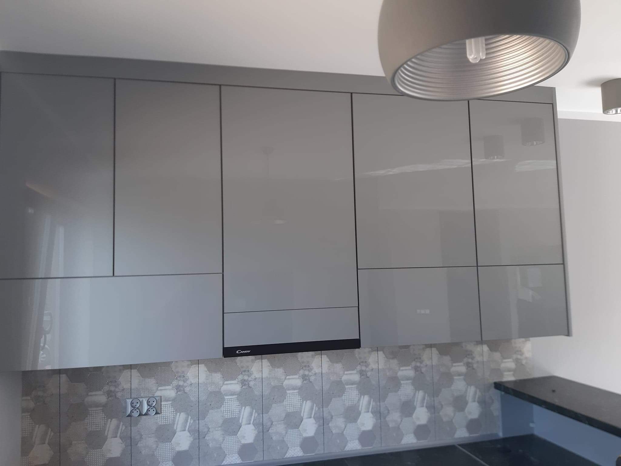 Szare, błyszczące szafki kuchenne z czarnym okapem marki Candy, heksagonalne płytki na ścianie, widoczne gniazdka elektryczne i lampa sufitowa.