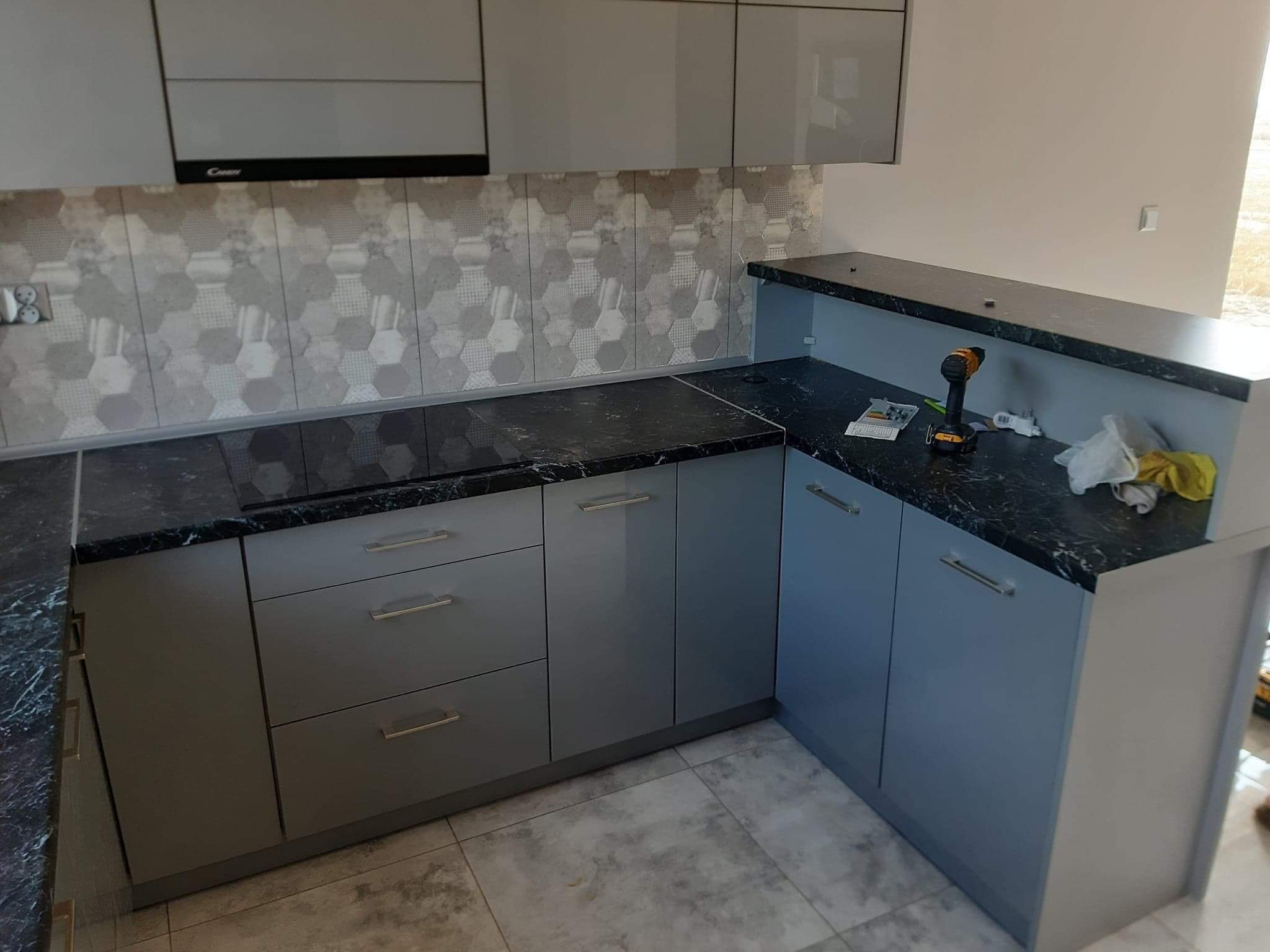 Kątowe ujęcie fragmentu szarej kuchni z czarnym blatem, widoczna płyta indukcyjna, szara mozaika na ścianie, wiertarka i akcesoria na blacie, prawdopodobnie w trakcie montażu.