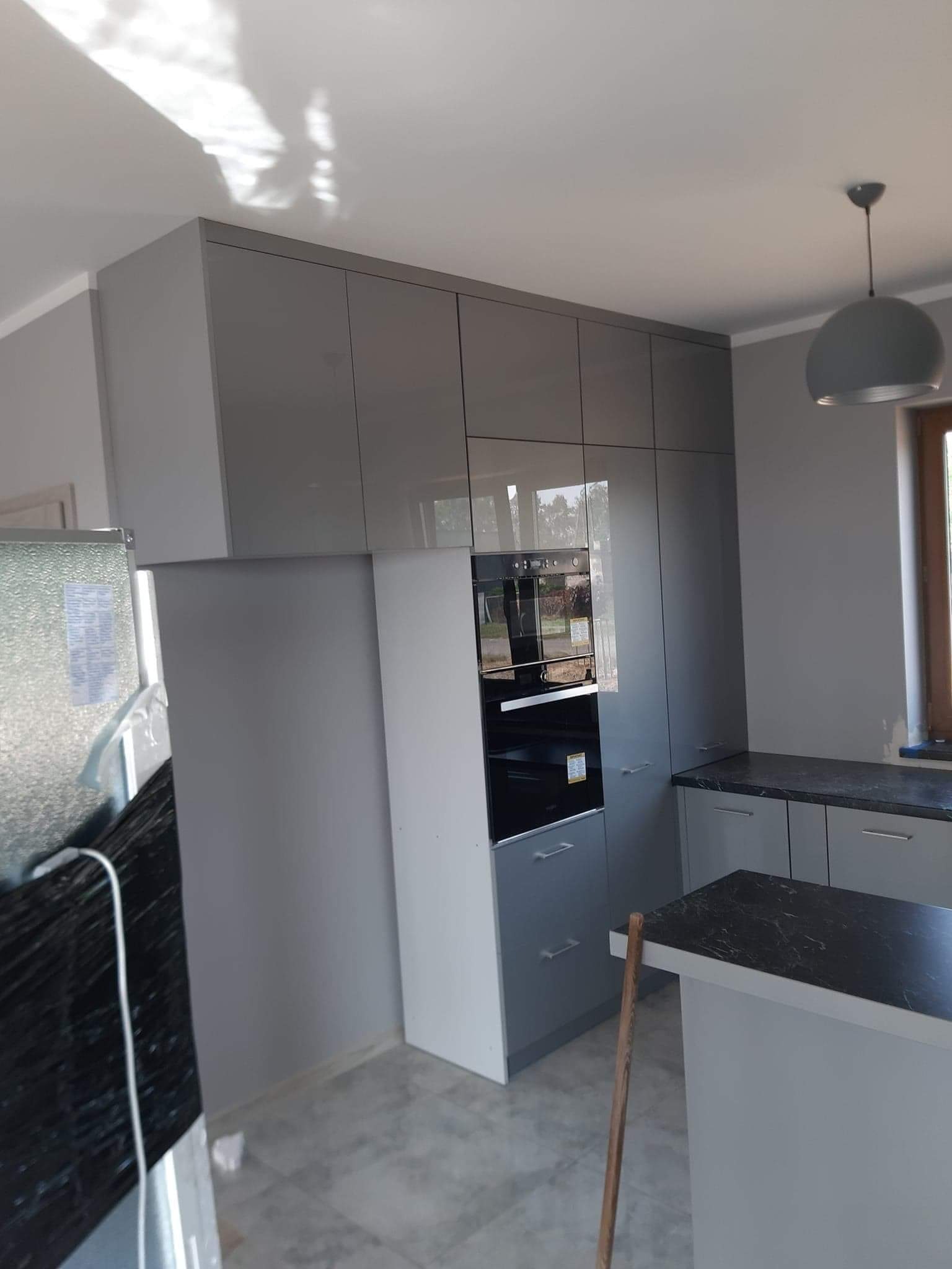 Szara kuchnia z zabudową pod sufit, piekarnik w zabudowie, wyspa kuchenna z czarnym blatem, widoczne elementy wykończeniowe.