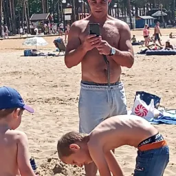 Mężczyzna w czapce robi zdjęcie telefonem na plaży, w tle widać drzewa i inne osoby.