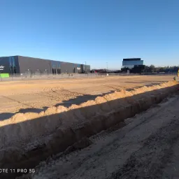 Żółta mini koparka wykonuje wykop liniowy w piaszczystym terenie budowy, z widocznymi śladami opon i budynkami przemysłowymi w tle, pod błękitnym niebem.