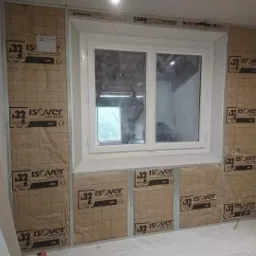 Ściana z oknem w trakcie ocieplania, widoczne płyty izolacyjne Isover, surowy stan wykończenia, przygotowanie do dalszych prac.