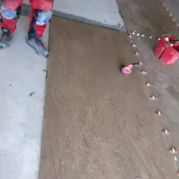 Układanie płytek podłogowych o wzorze kamienia, widoczne narzędzia glazurnicze i fragment roboczego ubrania wykonawcy.