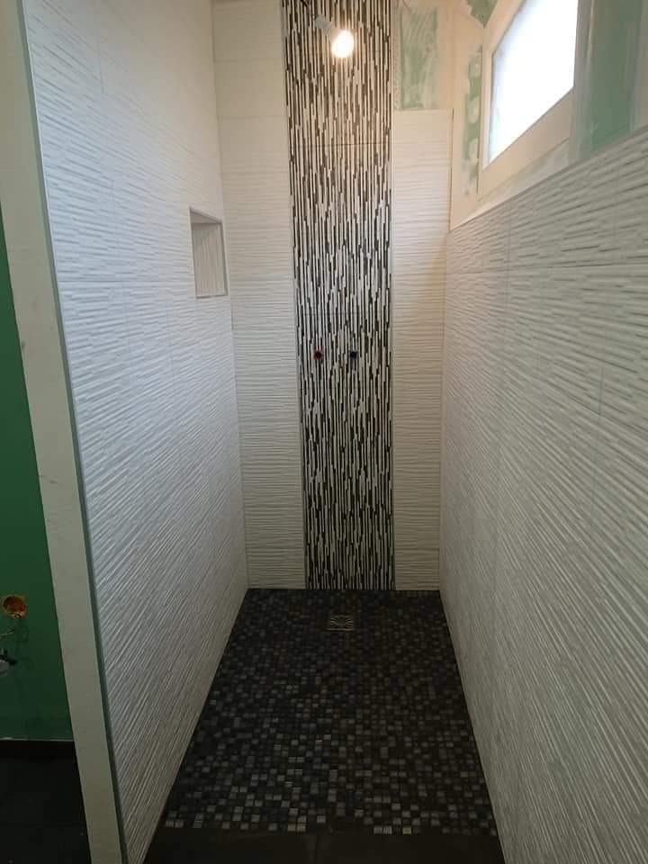 Wąska kabina prysznicowa z czarną mozaiką na podłodze i dekoracyjnym pasem płytek na ścianie, wykończona białymi płytkami z poziomym wzorem, widoczne elementy instalacji wodnej.