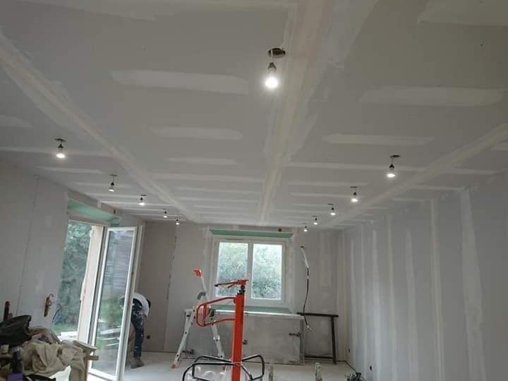 Pokój w trakcie prac wykończeniowych: płyty gipsowo-kartonowe na ścianach i suficie, zainstalowane punktowe oświetlenie sufitowe, widoczne narzędzia malarskie i drabina.