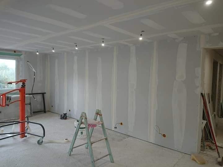 Wnętrze w trakcie prac wykończeniowych: ściany pokryte szpachlą, widoczne łączenia płyt gipsowo-kartonowych, drabina malarska, podnośnik do płyt i punkty oświetleniowe w suficie.