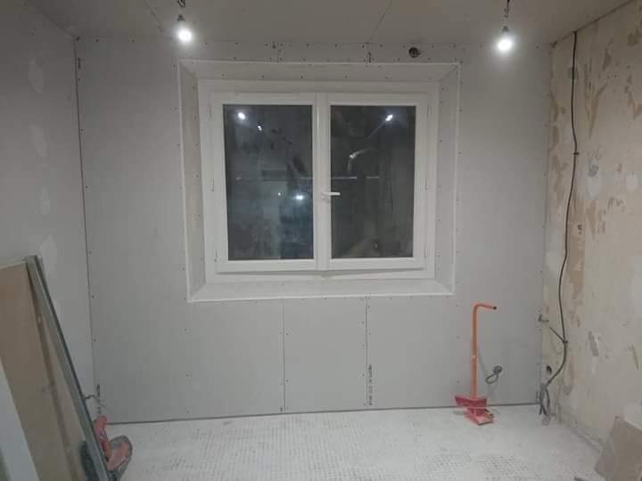 Pokój w trakcie remontu: ściany wyłożone płytami gipsowo-kartonowymi, widoczne łączenia i wkręty, okno w białej ramie, fragment nieotynkowanej ściany z instalacją elektryczną, narzędzia do obróbki...