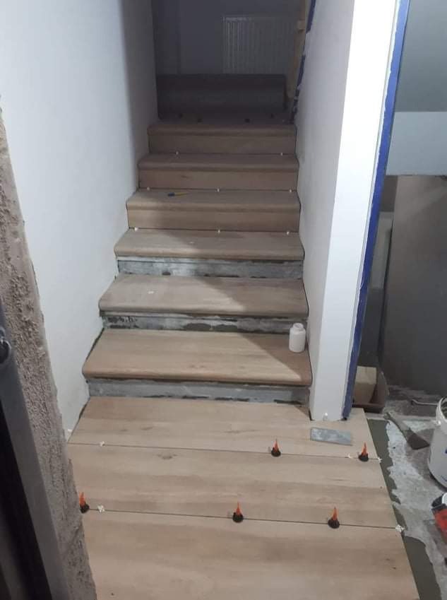 Montaż drewnopodobnych paneli na schodach, widoczne plastikowe krzyżyki dystansowe, butelka kleju na stopniu, surowe ściany w tle.