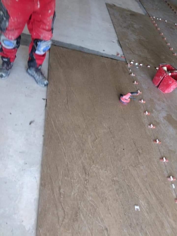 Układanie płytek podłogowych o wzorze kamienia, widoczne narzędzia glazurnicze i fragment roboczego ubrania wykonawcy.