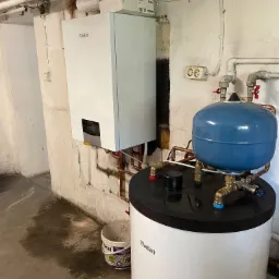 Biały piec gazowy Vaillant zawieszony na ścianie w piwnicy, obok biały zbiornik z niebieskim naczyniem wzbiorczym, widoczne rury miedziane i plamy wody na podłodze.