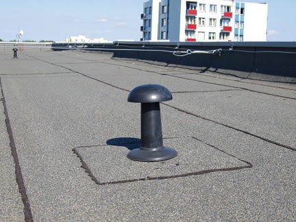 Czarny kominek wentylacyjny na pokrytym papą dachu płaskim budynku mieszkalnego, widoczne łączenia papy, jasne niebo w tle.