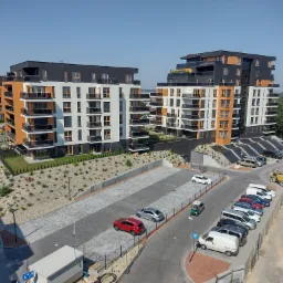 Nowoczesne osiedle z budynkami wielorodzinnymi o elewacjach w kolorach bieli, grafitu i drewna, z balkonami i parkingiem dla samochodów osobowych oraz zielenią wokół.