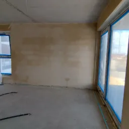 Wnętrze w stanie deweloperskim z nałożonymi tynkami, widoczne okna z niebieską folią ochronną, wylewka na podłodze i materiały budowlane w rogu pomieszczenia.