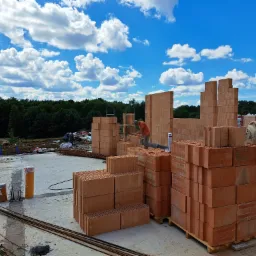 Wznoszenie ścian z pustaków ceramicznych na placu budowy; widoczne ułożone palety z materiałem, zbrojenia i pracownik w pomarańczowej koszulce na tle lasu i błękitnego nieba z chmurami.