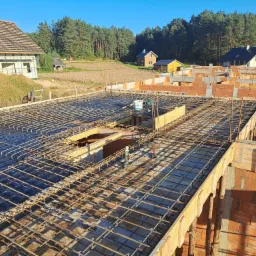 Zbrojenie stropu domu w trakcie budowy, widoczne pustaki ceramiczne, w tle inne budynki i las.