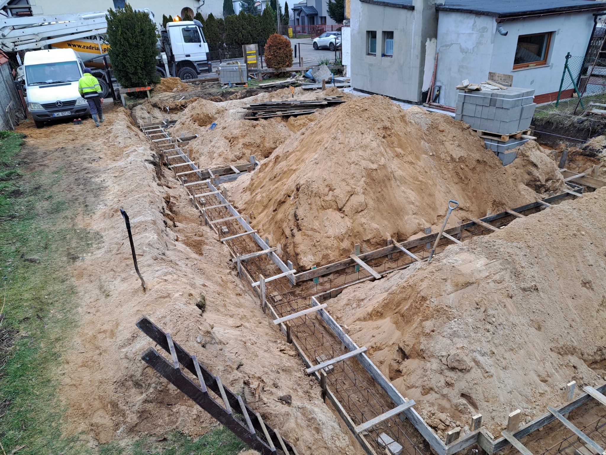 Wykop pod fundamenty z deskowaniem i zbrojeniem, widoczny stos bloczków betonowych, kopiec piasku oraz pracownik w odblaskowej kurtce w tle.