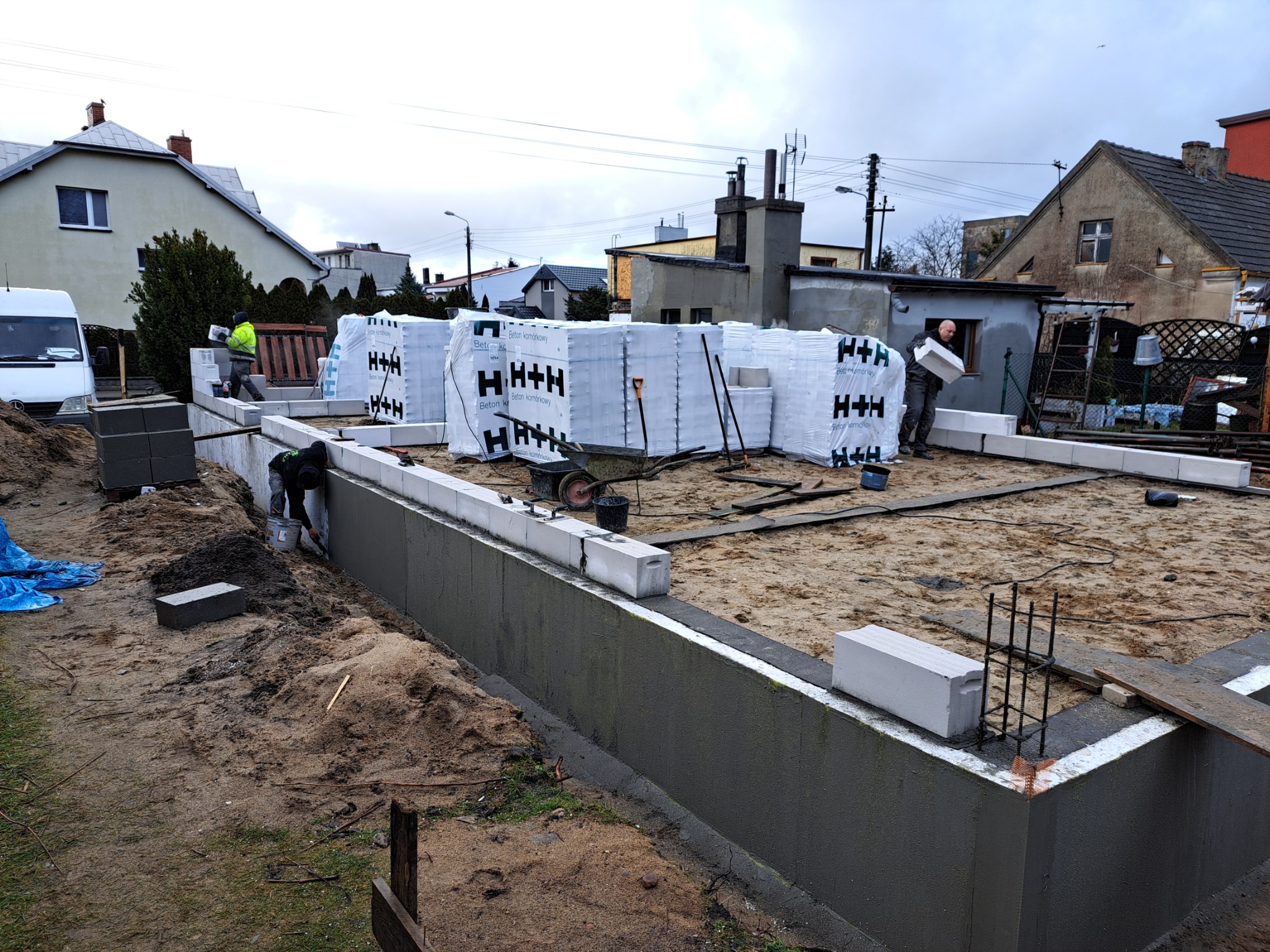 Budowa fundamentów: pracownicy układają bloczki betonowe H+H na przygotowanym fundamencie, widoczne zbrojenia, worki z bloczkami w tle, pochmurne niebo.
