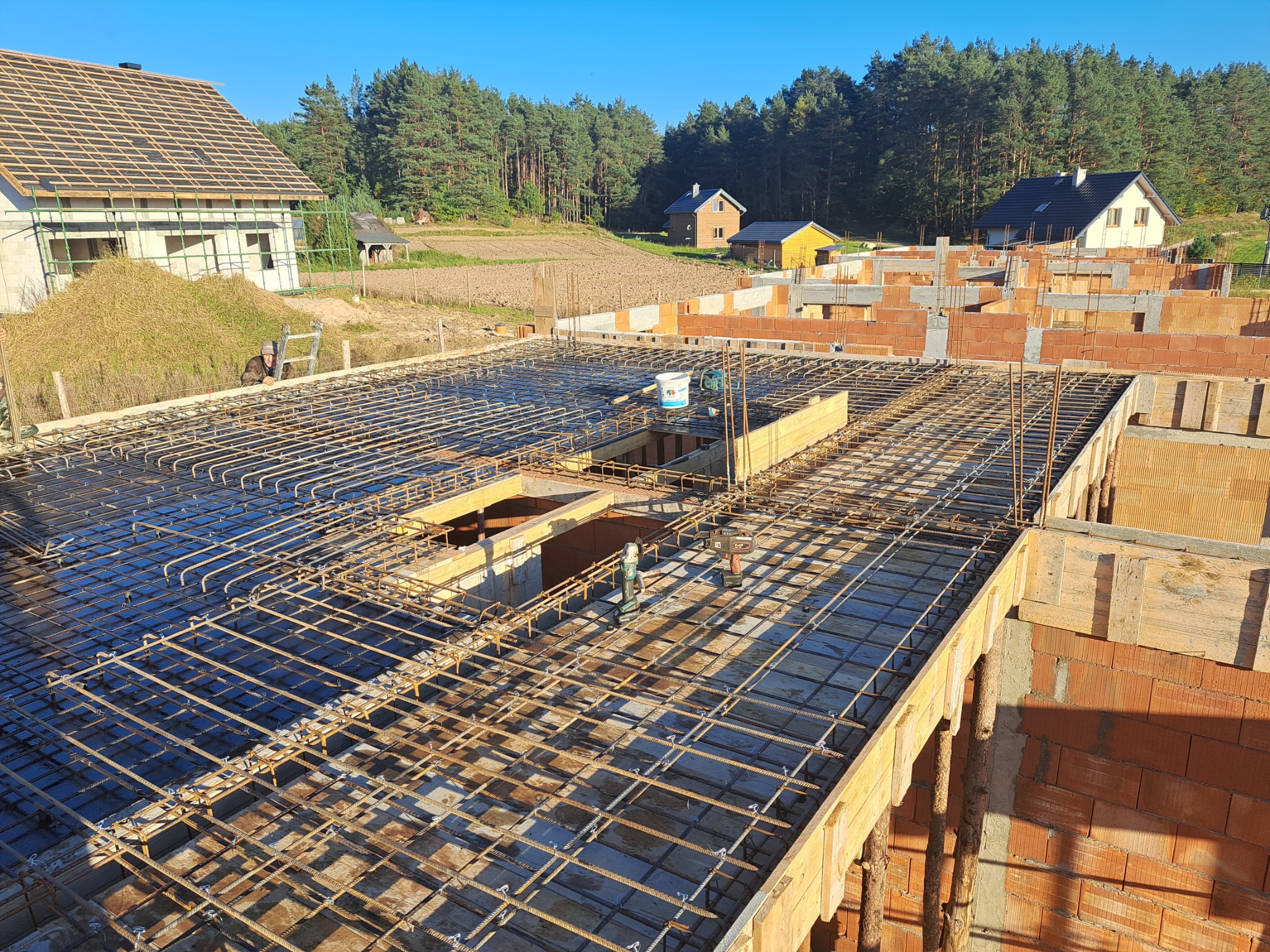 Zbrojenie stropu domu w trakcie budowy, widoczne pustaki ceramiczne, w tle inne budynki i las.