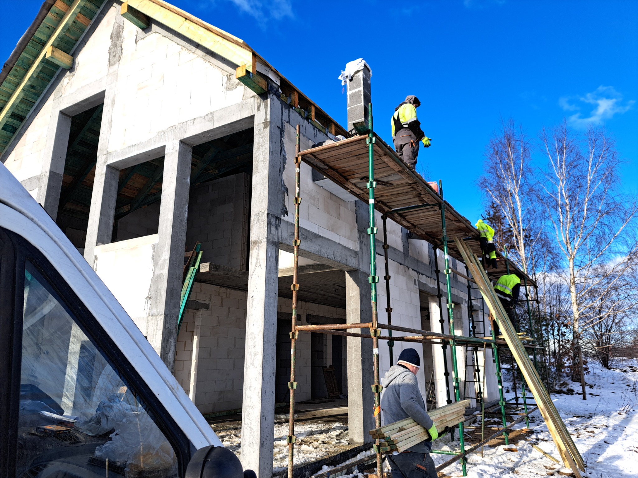 Budowa domu z pustaków: robotnicy na rusztowaniu w trakcie prac murarskich, widoczny fragment ośnieżonego terenu i samochodu dostawczego.