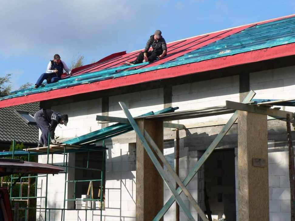 Budowa domu w Kniewie: Murarze układają czerwoną dachówkę na nowym budynku z białego bloczka, widoczne rusztowania i drewniane podpory.