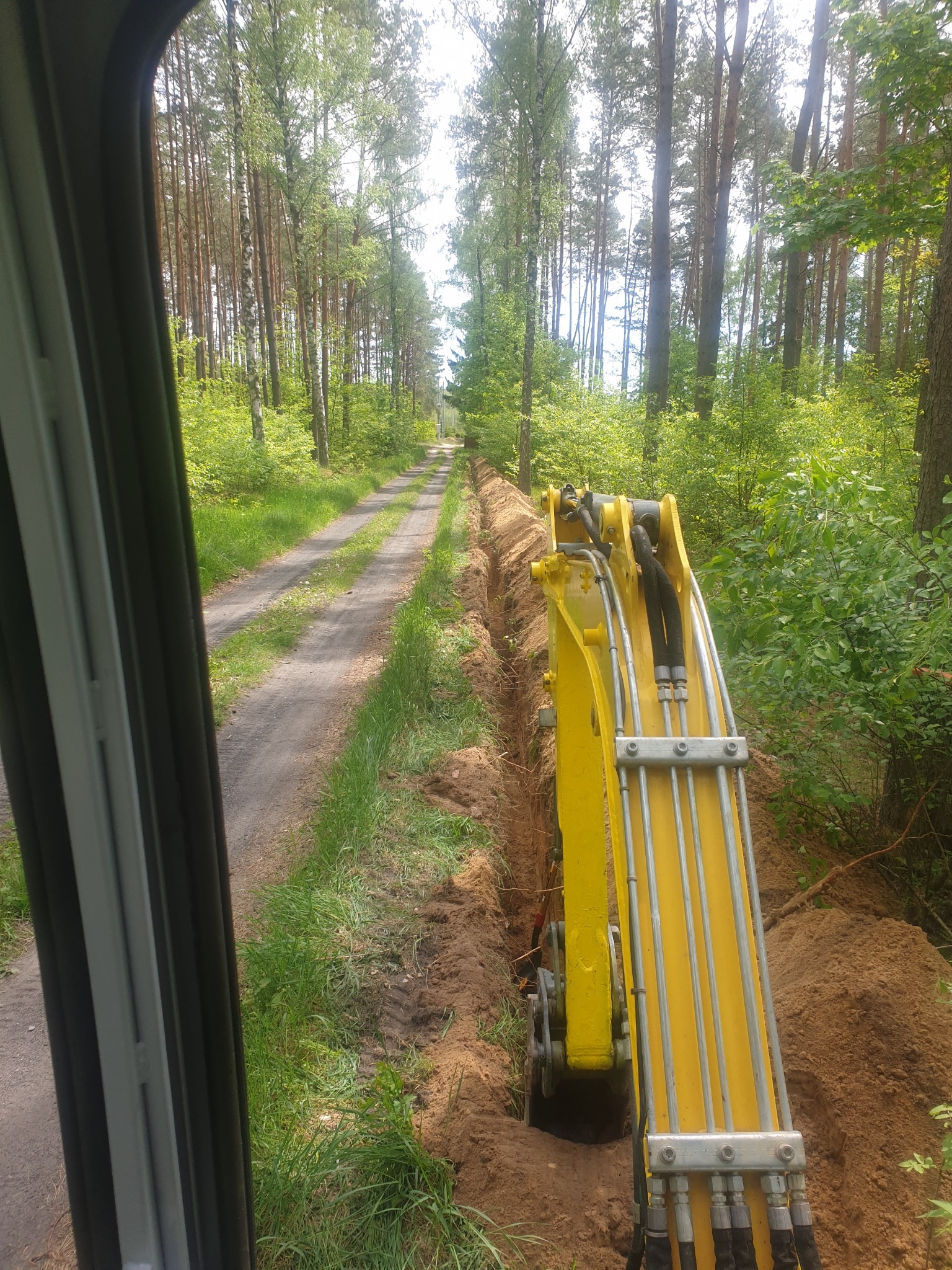 Żółta koparka w trakcie wykopywania wąskiego rowu w ziemi wzdłuż leśnej drogi, widok z kabiny operatora.