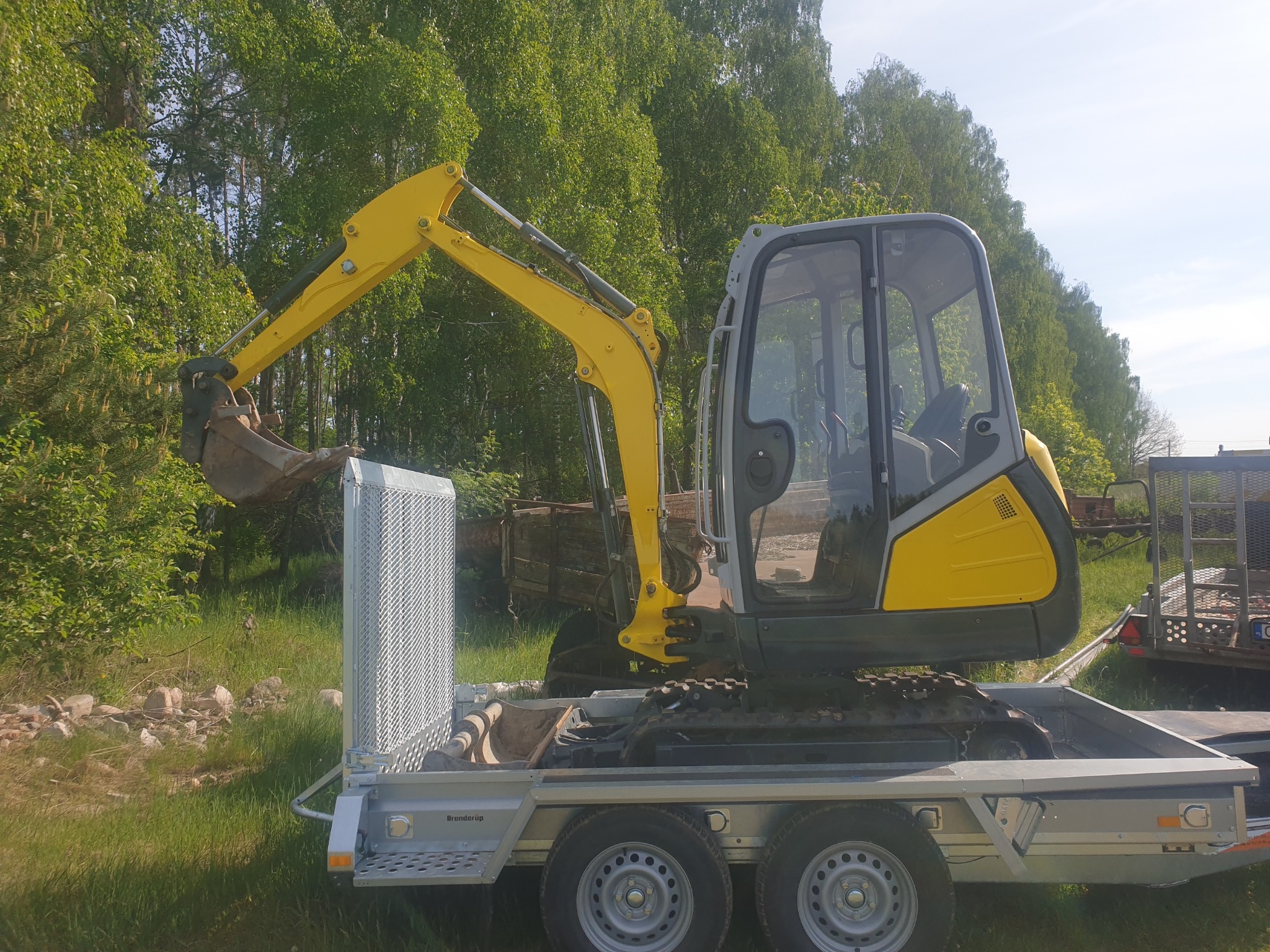 Żółto-szara mini koparka na przyczepie samochodowej z kratką ochronną, na tle zieleni drzew.
