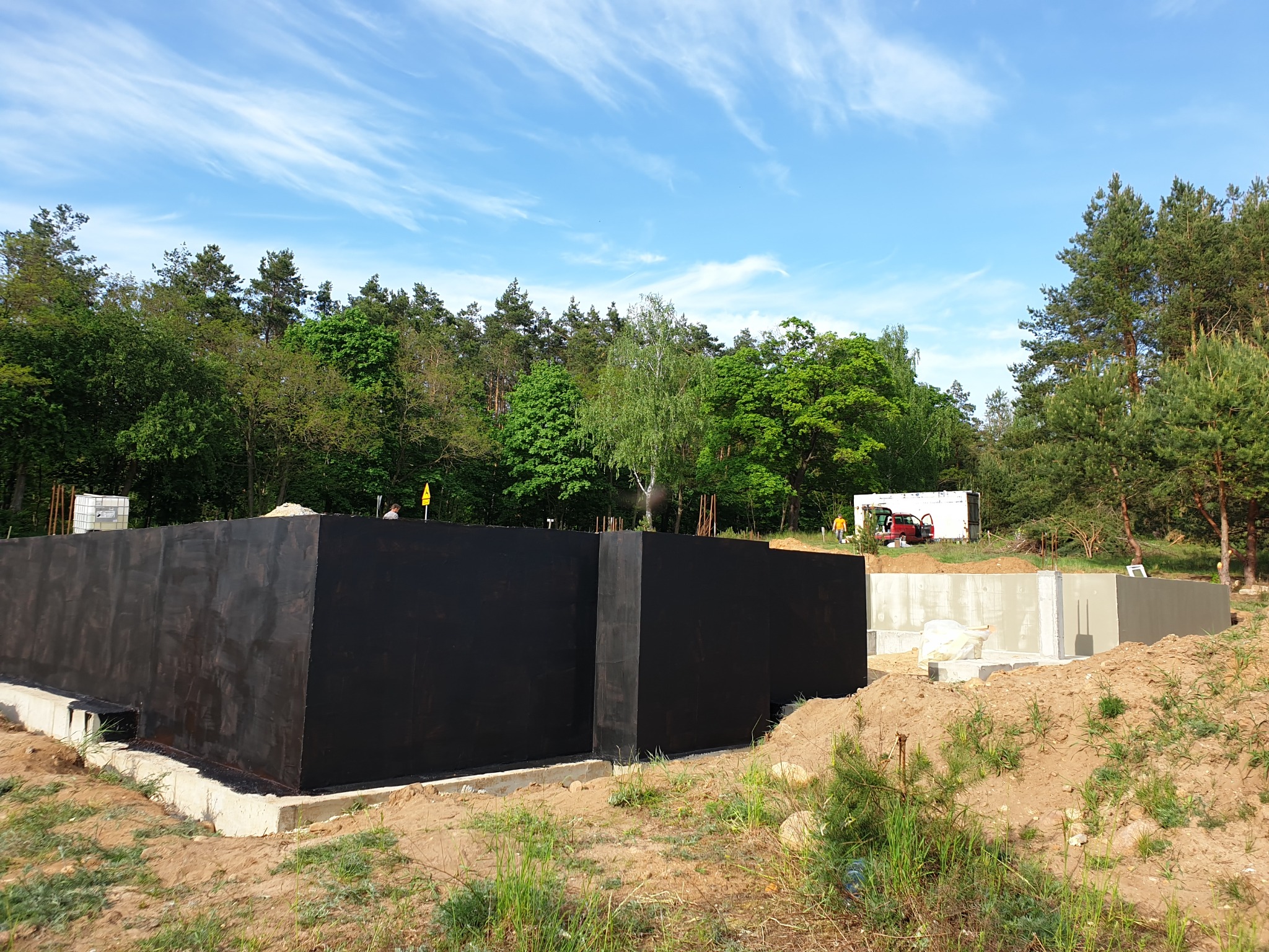 Zabezpieczone fundamenty budynku w trakcie budowy, widoczna czarna hydroizolacja, betonowe ściany fundamentowe, ziemia i zieleń w tle.