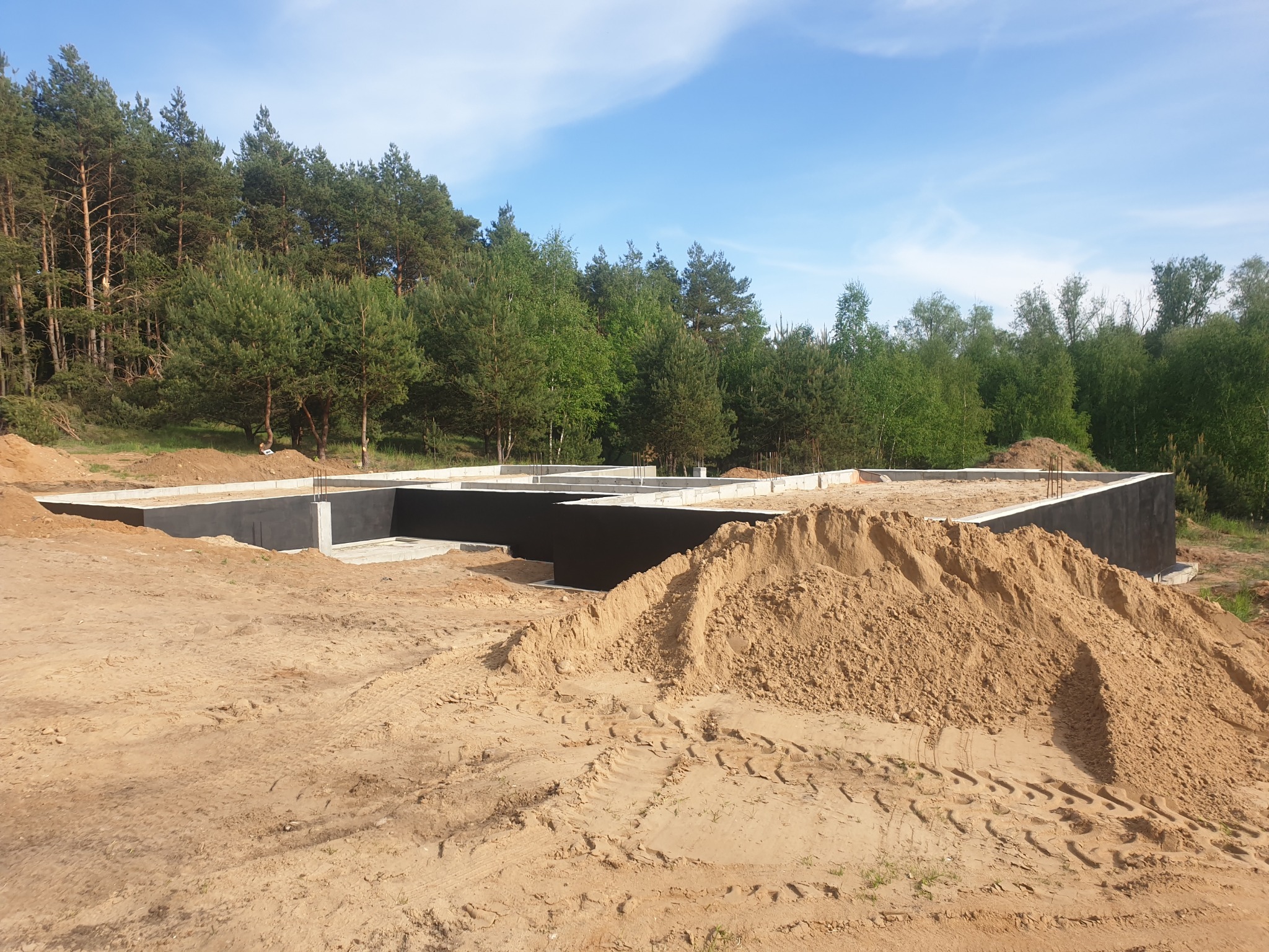 Fundamenty domu jednorodzinnego w trakcie budowy, z widoczną izolacją przeciwwilgociową i nasypaną ziemią, w tle las.