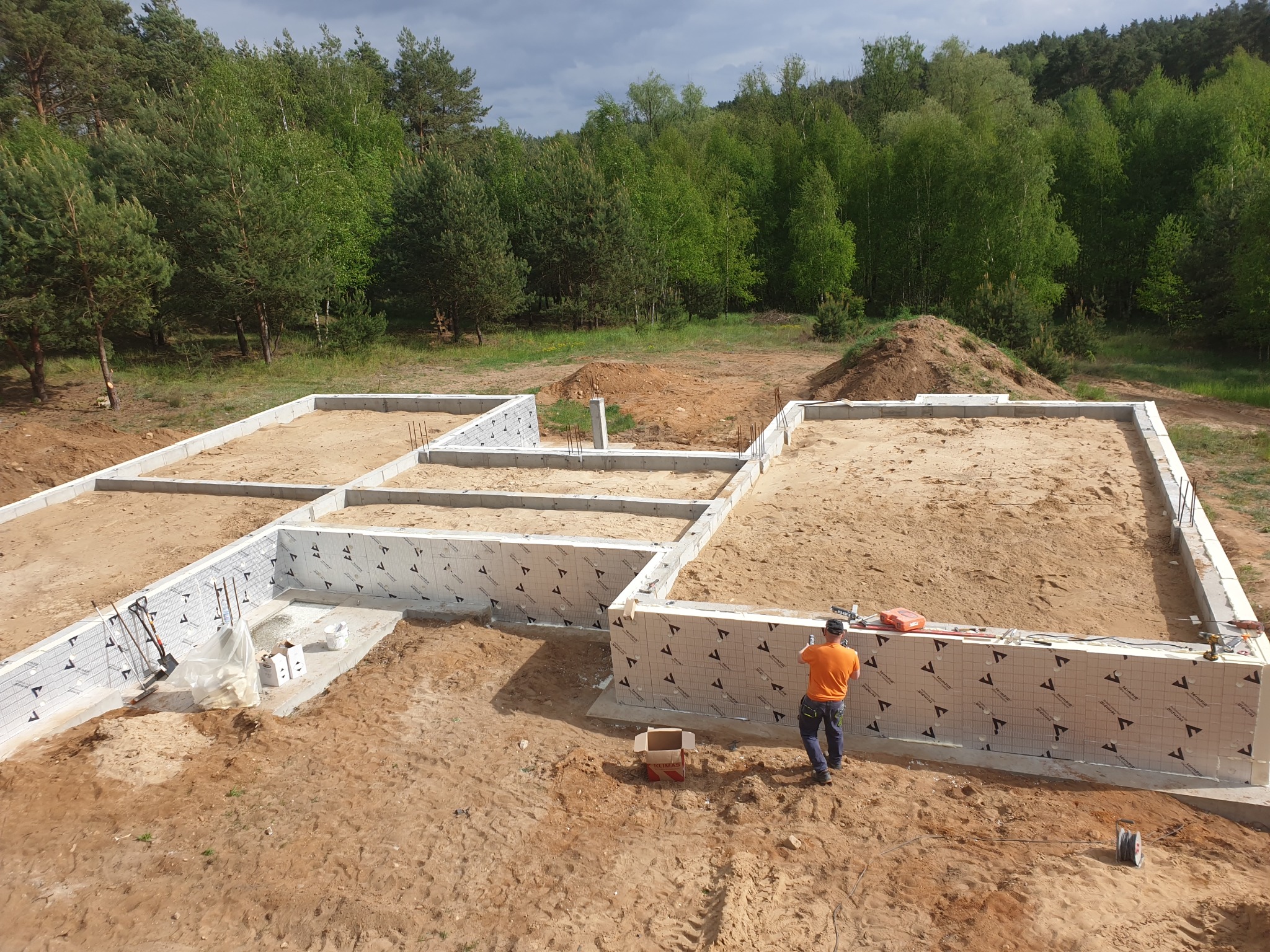 Budowa fundamentów domu jednorodzinnego z izolacją termiczną, widok z góry na wykop i pracownika w pomarańczowej koszulce przy ścianie fundamentowej na tle drzew