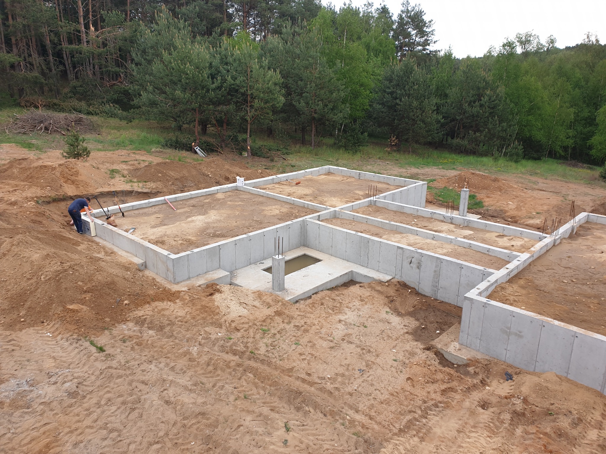 Wysoki kąt widzenia na fundamenty domu w trakcie budowy, z widocznym pracownikiem w niebieskiej koszuli przy lewym rogu, teren wokół wykopu pokryty piaskiem i ziemią, w tle las.