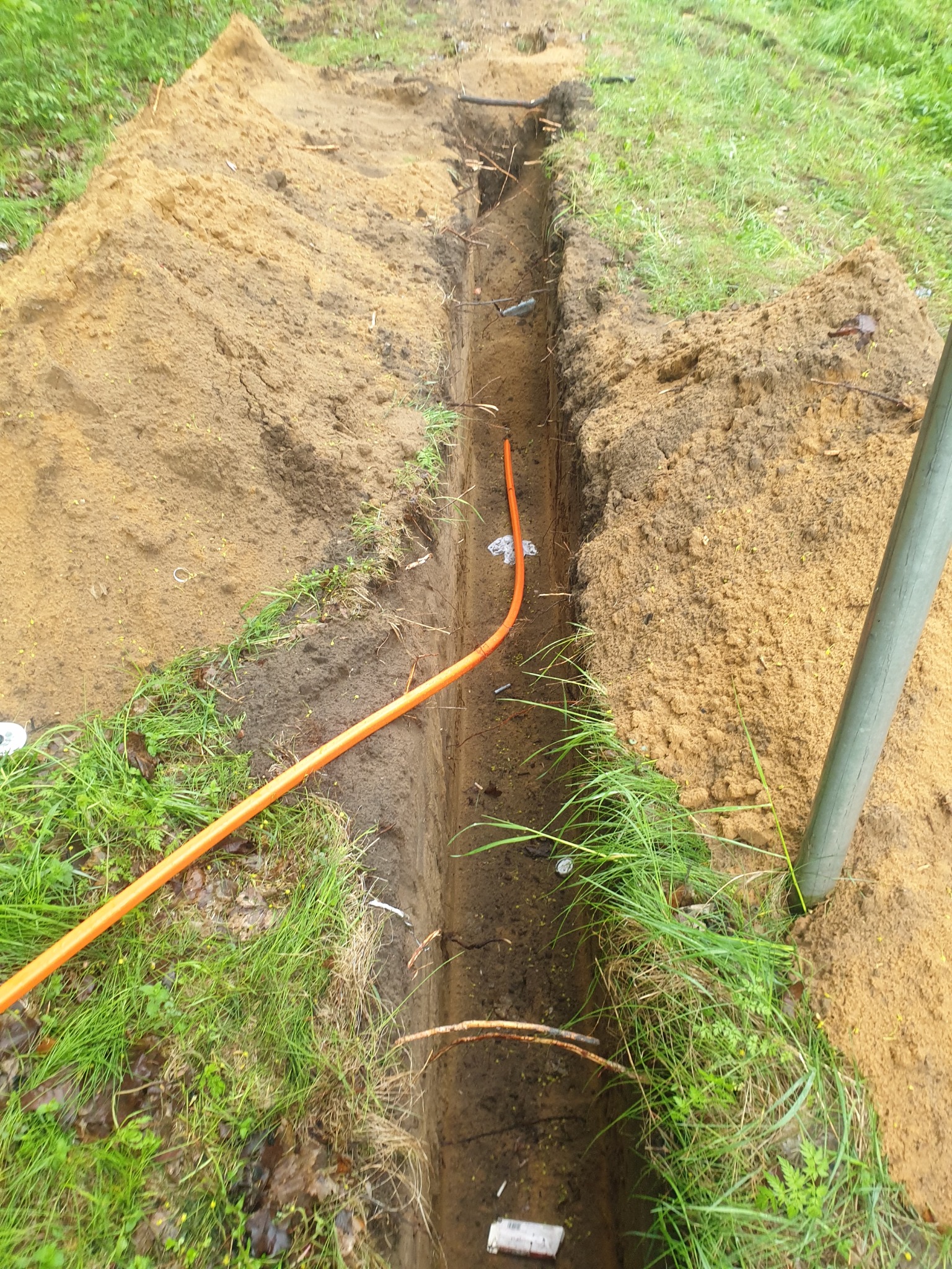 Wykopany wąski rów w ziemi z ułożonym pomarańczowym kablem, widoczne ślady wilgoci i świeżo naruszona gleba.