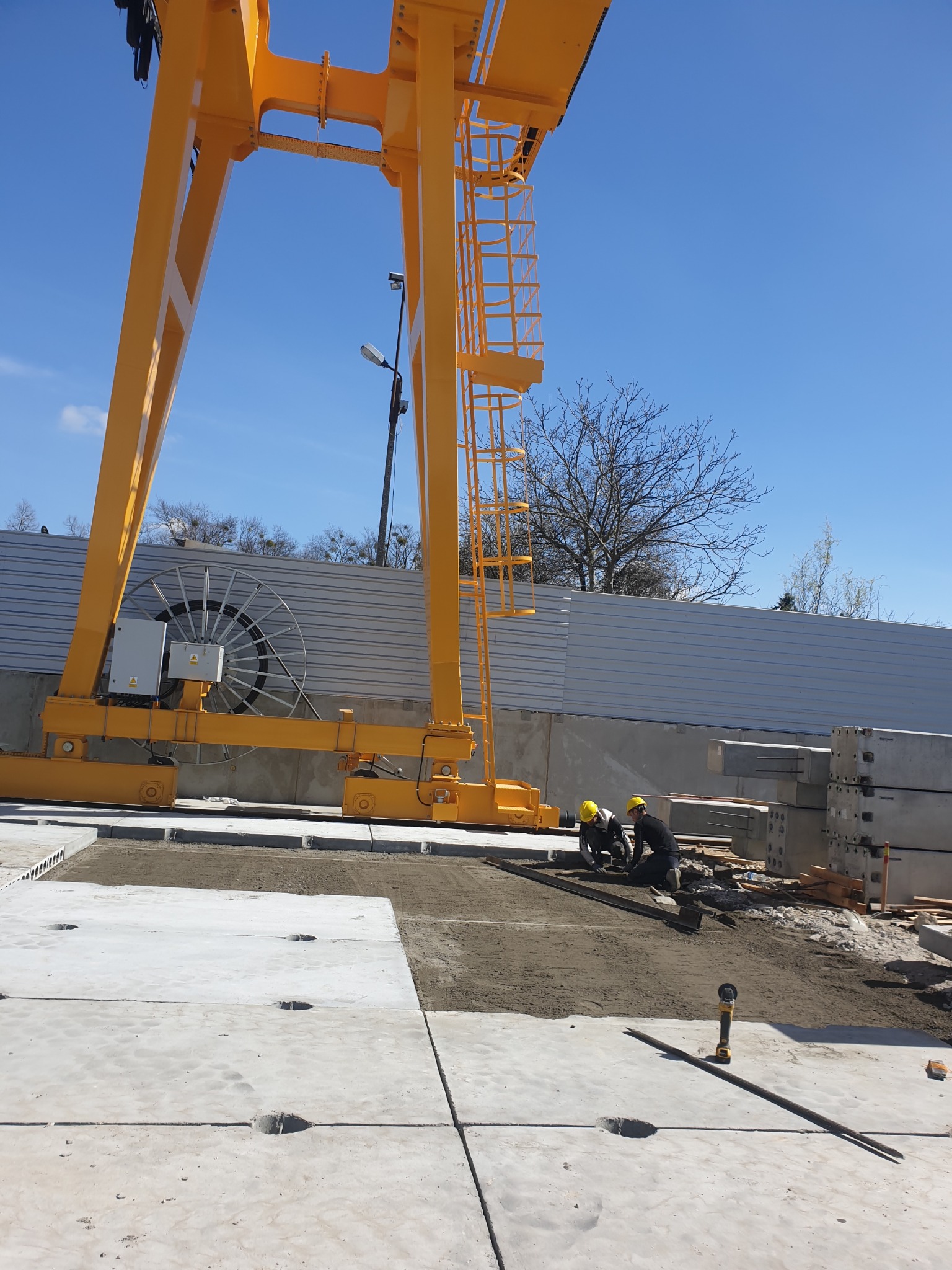 Montaż prefabrykowanych płyt betonowych pod suwnicą bramową na placu budowy, dwóch robotników w kaskach wyrównuje podłoże, widoczne zbrojenia i szlifierka kątowa.