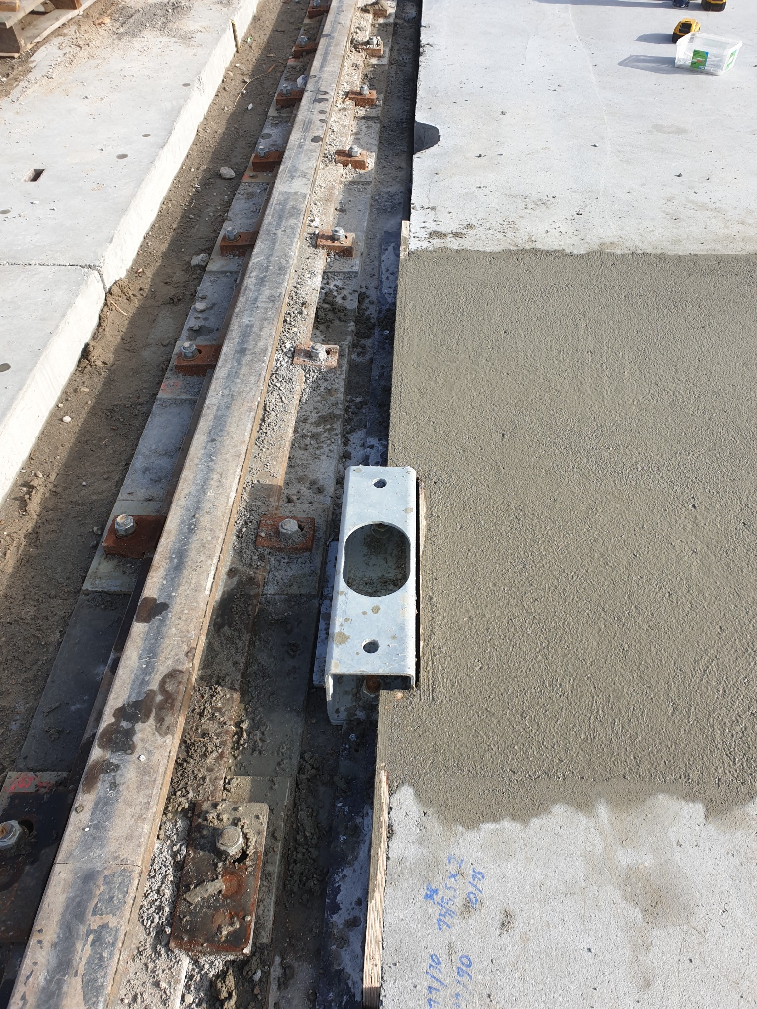 Świeżo wylany beton na placu budowy, widoczne metalowe szyny i mocowania, w tle narzędzia budowlane.