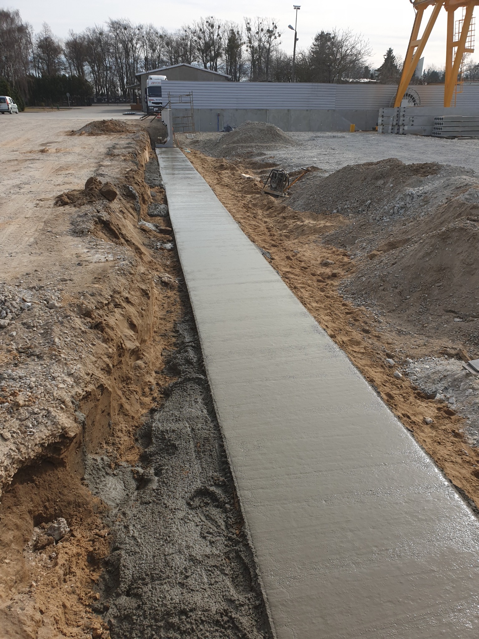 Świeżo wylany, długi, betonowy fundament w wykopie ziemnym, widoczny ubijak wibracyjny, w tle budynek i drzewa.