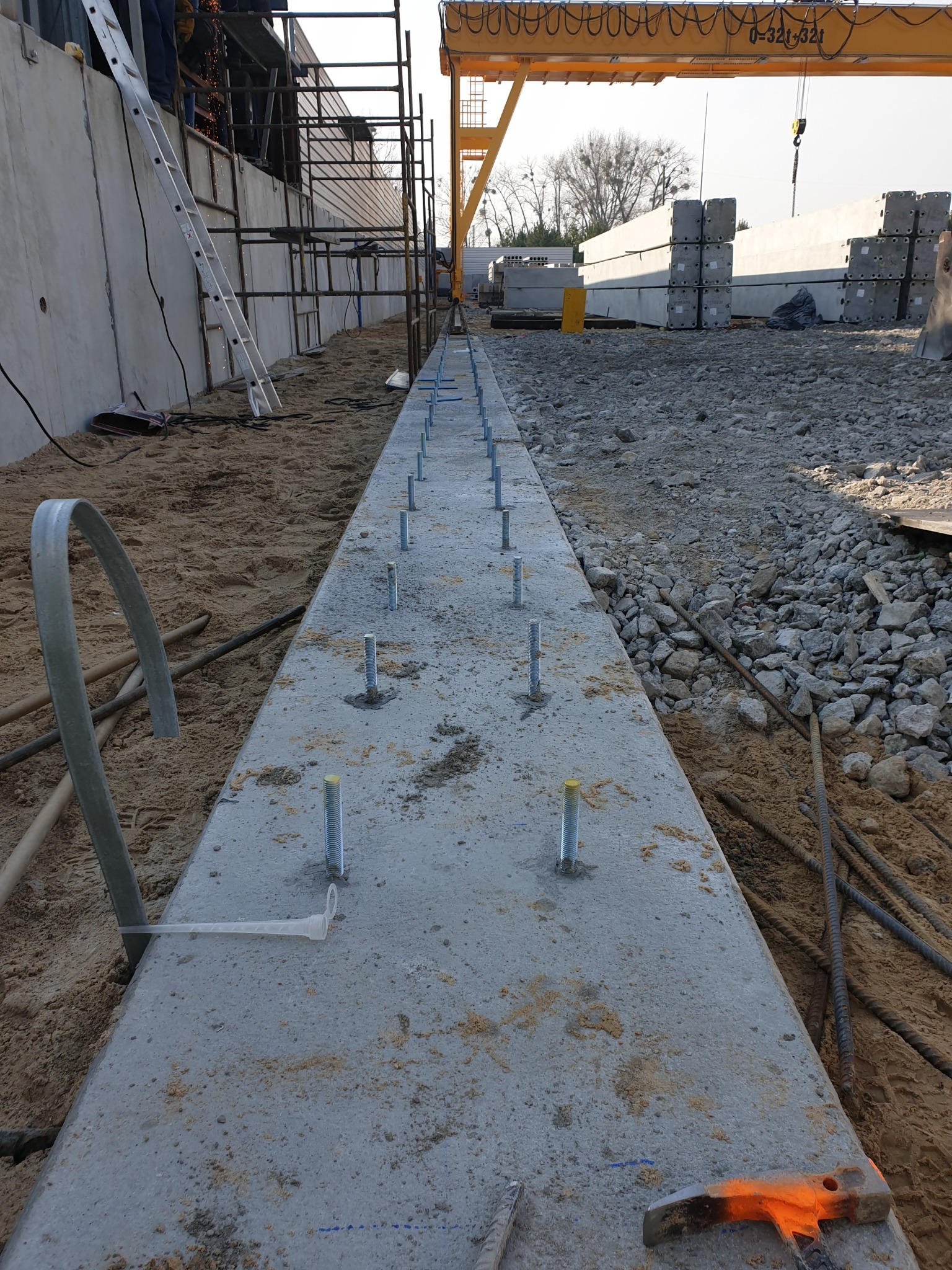 Wykop z przygotowanym podłożem betonowym z wystającymi śrubami mocującymi, widoczne rusztowania i dźwig w tle.