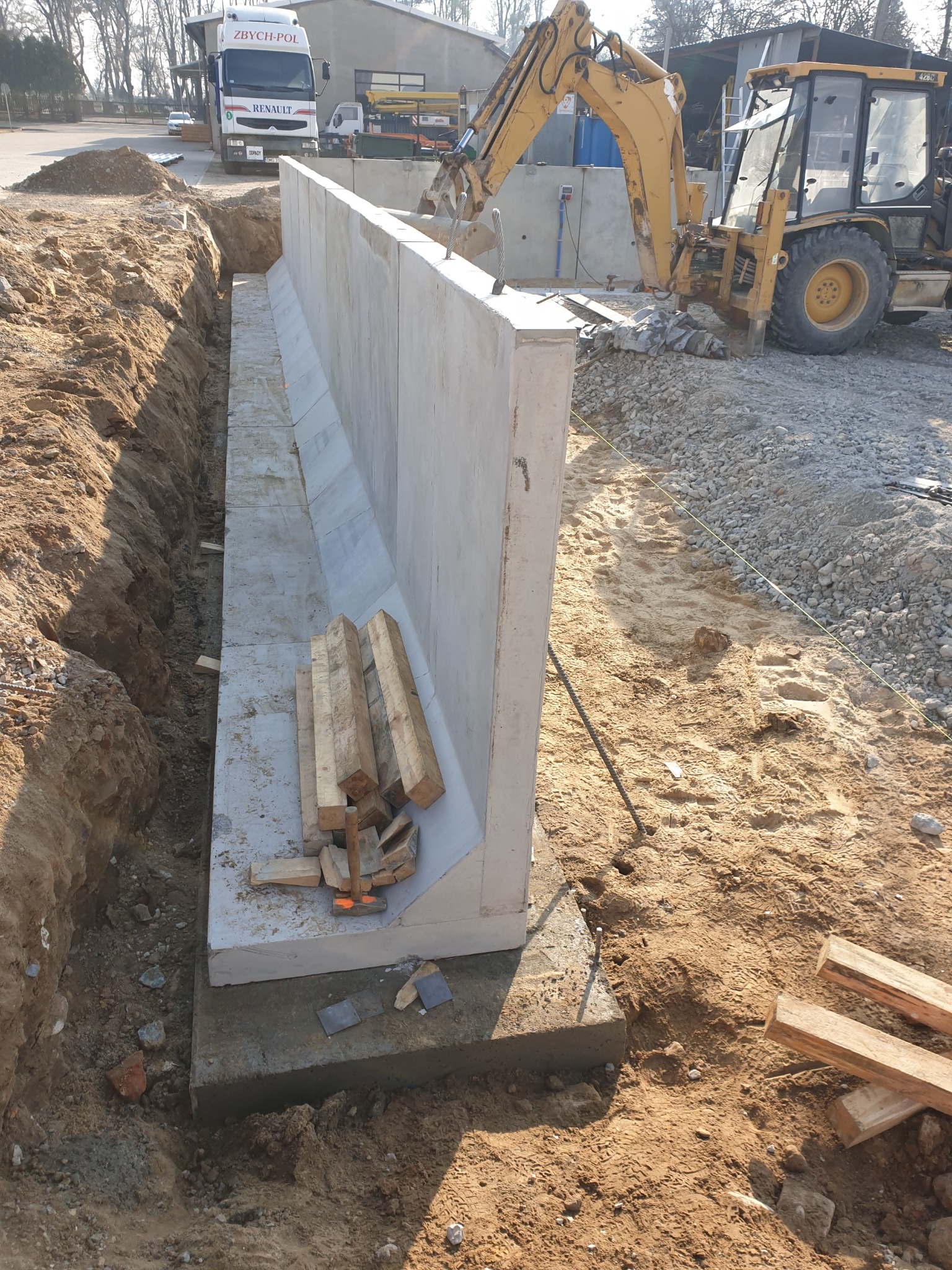 Wykop z betonową ścianą oporową w trakcie budowy; na pierwszym planie drewniane podpory, w tle koparka i ciężarówka ZBYCH-POL.